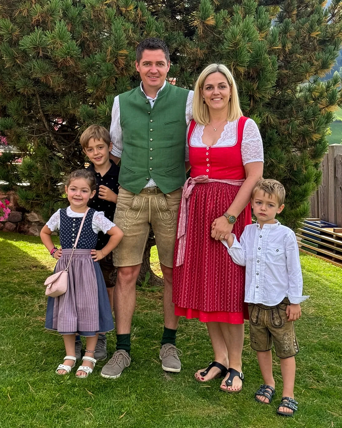 Familie Lesacher vom Sagritzer Wirt beim gemeinsamen Foto im Garten – Die Eltern, Gerald und Manuela Lesacher, in traditioneller Tracht mit ihren drei Kindern vor einem Nadelbaum.