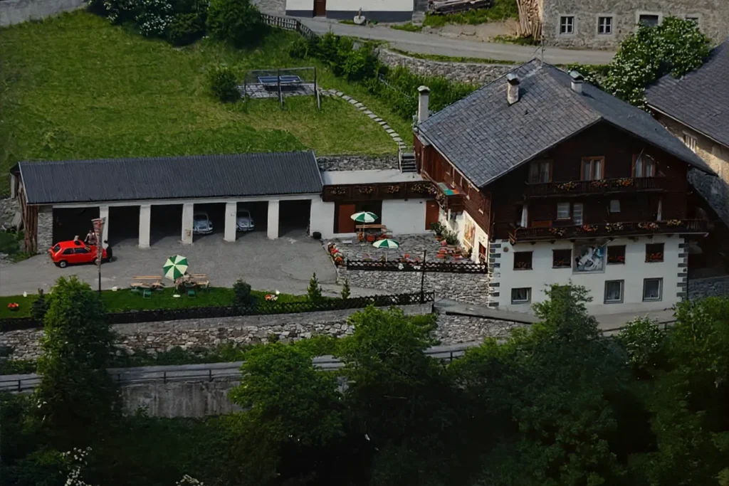 Historische Außenaufnahme des Sagritzer Wirts mit Garagen, Terrasse und dem markanten roten Auto im Hof, umgeben von grünen Wiesen und Bäumen.