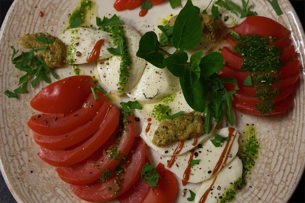 Frisch angerichtete Caprese-Variation mit Tomaten, Mozzarella, Kräutern, Pesto und dekorativen Toppings auf einem hellen Teller.